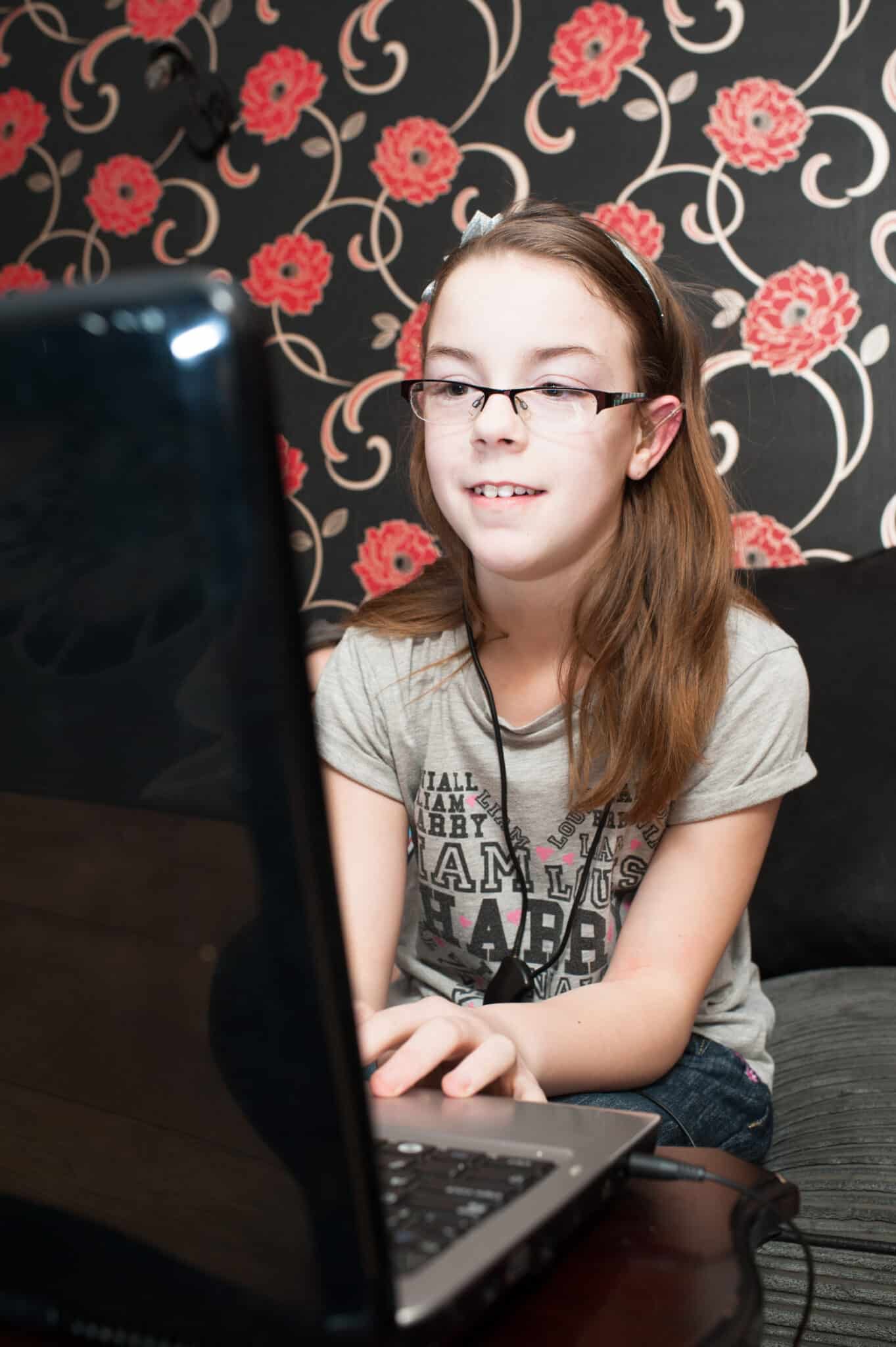 A girl with a hearing aid typing on a computer against a floral-print black wall